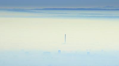 A single smokestack rises above a dense layer of fog covering an urban landscape, with distant mountains visible in the background.