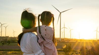 A woman with a young girl in her arms stands outdoors, looking at wind turbines during sunset.