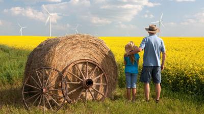 A man and a child stand in a field of yellow flowers next to a large hay bale on wheels, overlooking wind turbines in the distance.