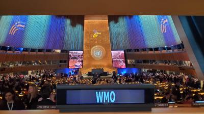 A large conference hall filled with attendees, featuring a stage with a large emblem and multiple screens displaying images. The foreground shows a sign with "WMO.