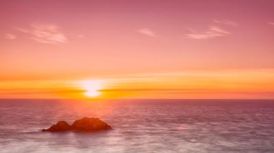 A serene sunset over a calm ocean with two small rocks in the foreground, under a pink and orange sky.