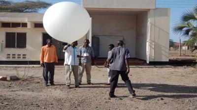 A group of people prepares to launch a large white weather balloon outside a small building on a sunny day.