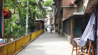 Narrow alley with brick buildings on one side, trees on the other, a yellow railing, and a person walking away in the distance.