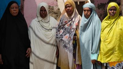 Five women stand side by side, wearing various colored hijabs. The background features a pink wall and a staircase.