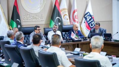 A group of people are seated around a conference table in a formal meeting room, with multiple flags and logos displayed on the walls.