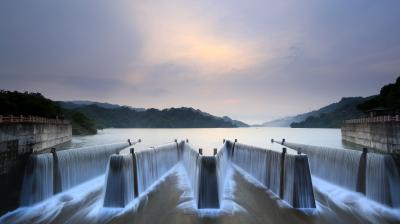 A symmetrical dam with flowing waterfalls under a cloudy sky, surrounded by lush green hills and a tranquil lake.