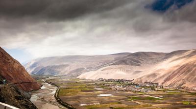 A scenic view of a river winding through agricultural fields and a small settlement, surrounded by rolling hills under a cloudy sky.