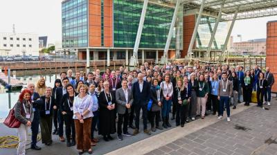 A large group of people pose for a photo outside a modern building with glass and brick elements. They stand on a paved area, some wearing lanyards. The setting appears to be a professional event.