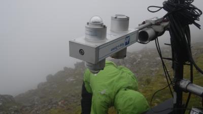 A person in a green jacket adjusts equipment on a foggy, rocky hilltop.