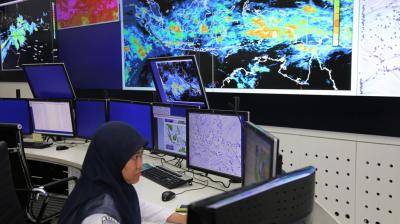 A person in a hijab works at a desk with multiple monitors displaying weather data and maps in a control room. Large screens on the wall show satellite images.