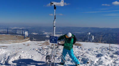 Person in winter gear adjusts a weather station on a snowy mountain summit under a clear blue sky.