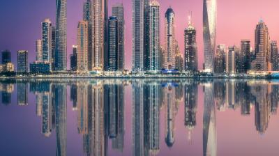 City skyline with tall, illuminated skyscrapers reflecting in a calm body of water during twilight. The sky is a gradient of pink and purple hues.