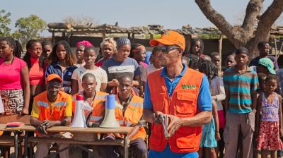 A group of people, including several in orange vests, stand outdoors near tables with megaphones. One person in an orange cap addresses the crowd.