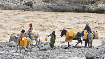 People and donkeys carrying yellow containers navigate a rocky riverbank with a fast-flowing river in the background.