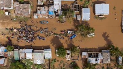 Aerial view of a flooded village with submerged buildings and boats. A large group of people gathers on dry ground, surrounded by muddy water. Trees and damaged structures are visible.