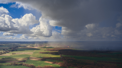 Aerial view of patchwork farmland under partly cloudy skies, with sunlight on one side and rain clouds casting shadows on the other.