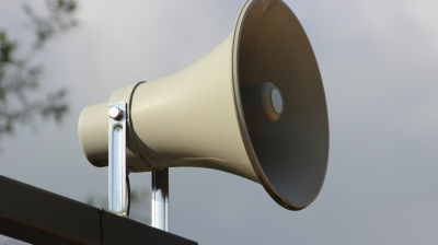 A close-up of a gray outdoor loudspeaker mounted on a metal bracket against a cloudy sky background.