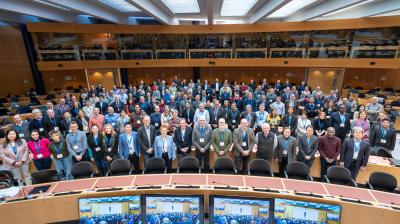 A large group of people pose for a group photo in a modern conference room with curved desks and computer monitors at the front.