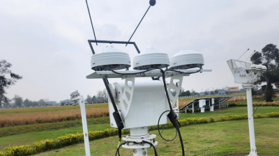 A weather monitoring instrument with three white sensor modules and antennae stands on a grassy field with crops and cloudy sky in the background.