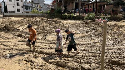 Three people walk through deep mud with building debris and tire tracks in an urban area, with residential houses and onlookers in the background.
