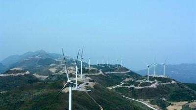 A series of wind turbines are installed along a hilly landscape, with winding roads running through the area under a clear sky.