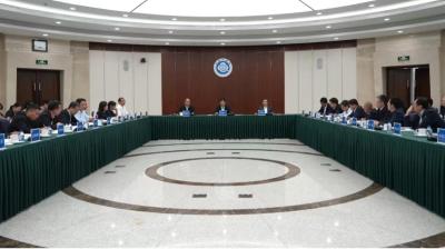 A formal meeting with over twenty people seated around a large U-shaped table in a conference room, with microphones and nameplates arranged in front of them.