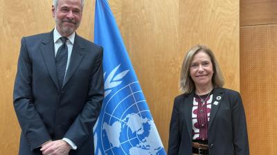 Two people in business attire stand next to a blue United Nations flag, posing for a photo against a wooden wall background.