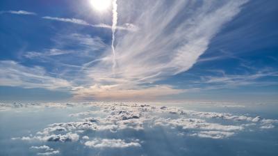 Sun shines brightly above a layer of scattered clouds, with streaks of white clouds and contrails stretching across a blue sky.
