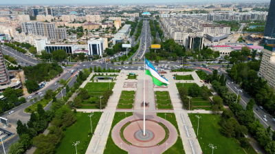 Aerial view of a city square with a large flagpole displaying the Uzbekistan flag, surrounded by green lawns, roads, and buildings.