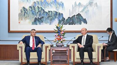 Two men in suits sit in armchairs facing each other with a flower arrangement between them, while a woman takes notes in the background. A large painting hangs on the wall behind them.