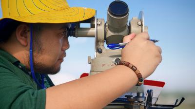 A person wearing a yellow hat looks through a surveying instrument while holding a pen and clipboard outdoors.