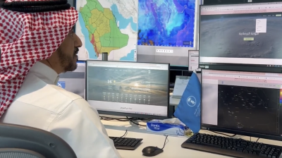 A man in traditional Middle Eastern attire monitors weather data and maps on multiple computer screens at a meteorological center.