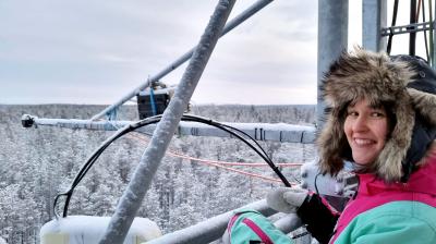 A person in a fur-lined hooded coat stands on a metal structure above a snowy forest, smiling at the camera.