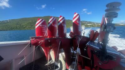 Several red and white Wave Glider marine drones are secured on the back of a boat, with the ocean and a green, hilly coastline visible in the background.