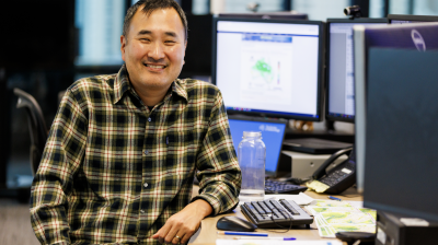 A man in a plaid shirt sits and smiles at a desk with multiple computer monitors and papers in an office setting.