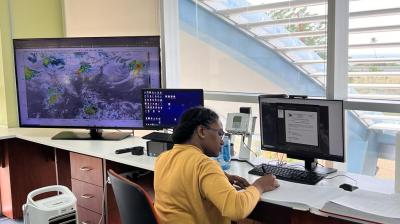A person in a yellow sweater works at a desk with two monitors; one displays a weather map, the other shows a document. A large window and portable air conditioner are visible.