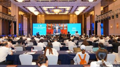 A large conference room filled with attendees facing a stage and screens displaying a speaker’s image during a formal event.