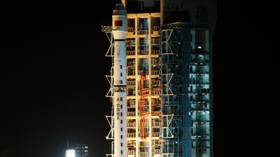 A rocket launches at night from a lit-up launch pad, emitting bright flames and smoke at the base against a dark sky.