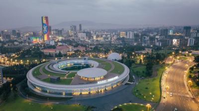 An aerial view of a modern, circular building with a central courtyard, surrounded by greenery and city lights, with a city skyline and tall buildings in the background at dusk.