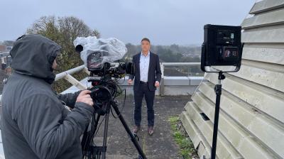 A man in a suit stands on a rooftop being filmed by a camera operator; professional lighting equipment is set up, and the weather appears overcast.