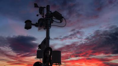A weather station with various sensors stands silhouetted against a dramatic sunset sky with red, orange, and blue clouds.