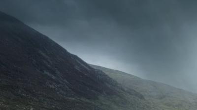 Dark, overcast sky above a sloping, rocky hillside with patches of grass and sparse vegetation.