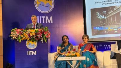 A man speaks at a podium decorated with flowers, with two seated women on stage beside him at an IITM scientific conference event. A presentation slide is visible in the background.