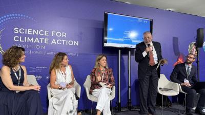 Five panelists sit and stand on a stage at the Science for Climate Action Pavilion, with a man speaking into a microphone and a presentation screen behind them.