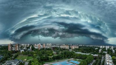 A dramatic, layered storm cloud formation looms over a cityscape with residential buildings and green spaces visible below.