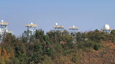 Several radar domes are mounted on tall metal towers above a forested hillside under a clear blue sky.