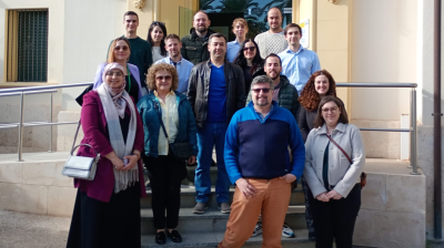 A group of sixteen adults stands on steps outside a building entrance, posing for a photo on a sunny day.