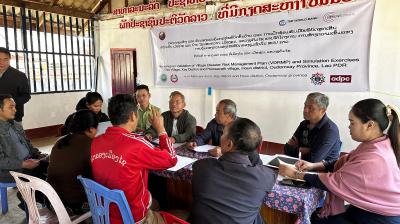 A group of people sit around tables in a meeting room, discussing documents. A banner about disaster risk management in Laos hangs on the wall behind them.