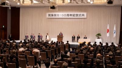 A formal ceremony in a large hall with seated attendees, a speaker at the podium, and banners in Japanese, alongside the Japanese flag and another flag.
