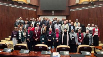 A group of people pose for a group photo inside a large conference room with wooden paneling and empty seats in the foreground.